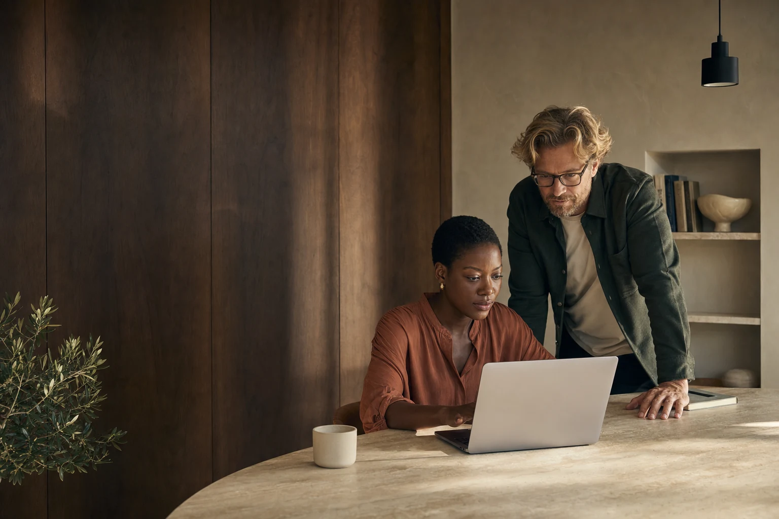 Two people reviewing work together on a laptop in a warmly lit studio with walnut walls.