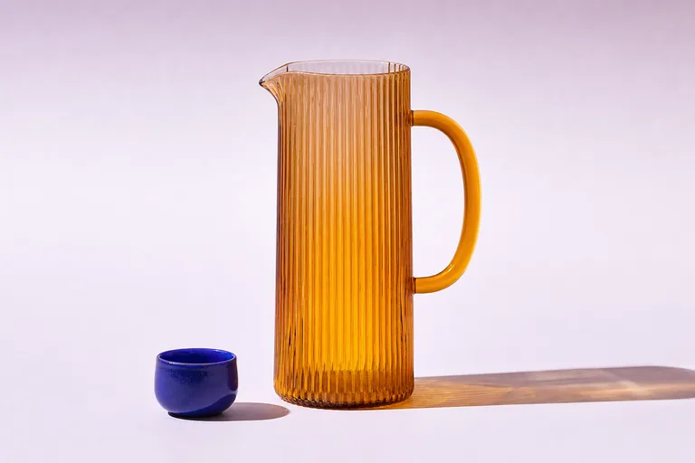 Illustrative product photograph of a ribbed amber-glass carafe on a pale lilac surface with a single cobalt-blue cup.