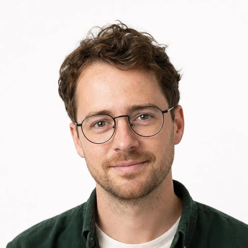 A man with wavy brown hair and round glasses, wearing a forest-green overshirt against a white background